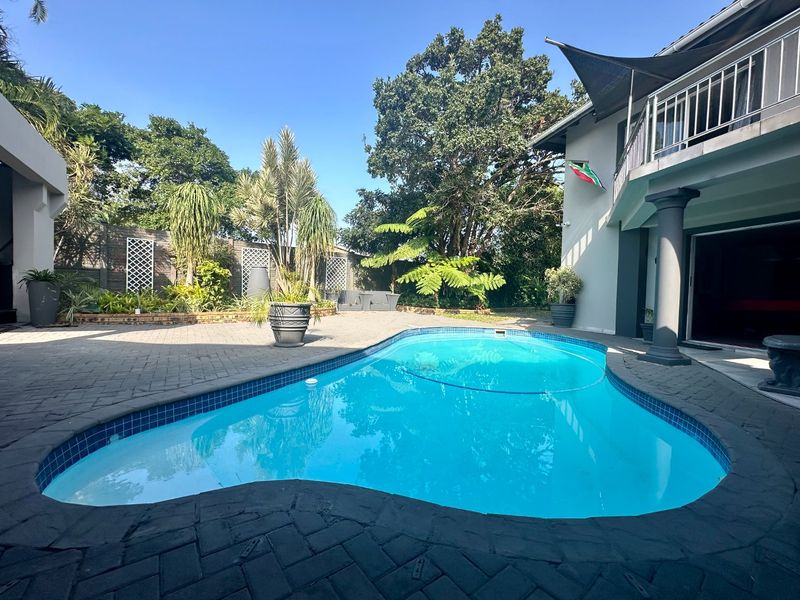 A well-maintained outdoor pool area featuring a paved patio, swimming pool, and a covered balcony with shade sail, set within a landscaped garden.