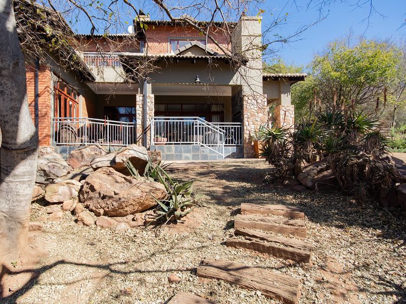 A rear view of the house featuring a covered patio with stone cladding, brick facade, and a landscaped garden with natural rock features.