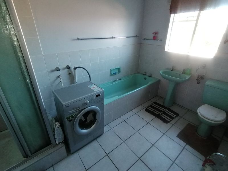 A dated bathroom featuring a pastel green bathtub, pedestal basin, and toilet, with tiled flooring and walls, currently accommodating a laundry appliance.
