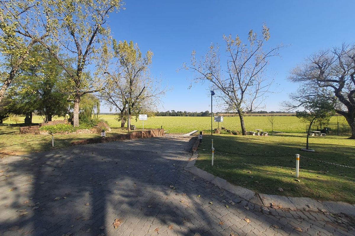 A paved driveway leading towards an open field, featuring mature trees and landscaped garden areas within the property grounds.