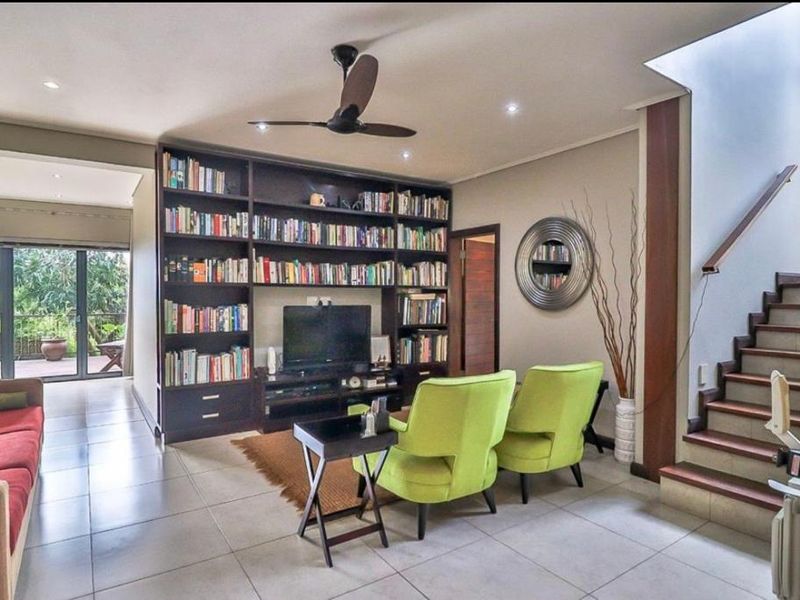 A spacious living room featuring a built-in floor-to-ceiling bookshelf, tiled flooring, a ceiling fan, and an adjacent staircase leading to the upper level.