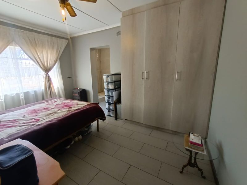 A bedroom featuring light wood-finish built-in cupboards, tiled flooring, and a ceiling fan, with direct access to an en-suite bathroom.