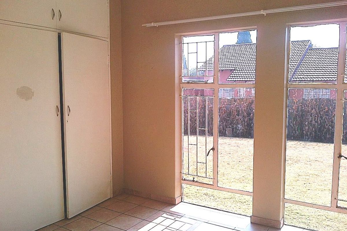 A vacant bedroom featuring built-in cupboards, tiled flooring, and large steel-framed windows with security bars overlooking the garden.