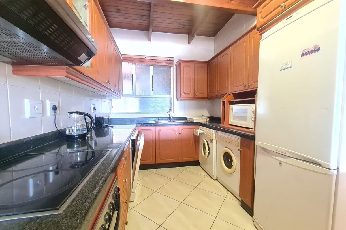 A functional kitchen featuring wooden cabinetry, dark granite countertops, tiled flooring, and exposed timber ceiling beams, equipped with built-in oven, hob, and extractor fan.