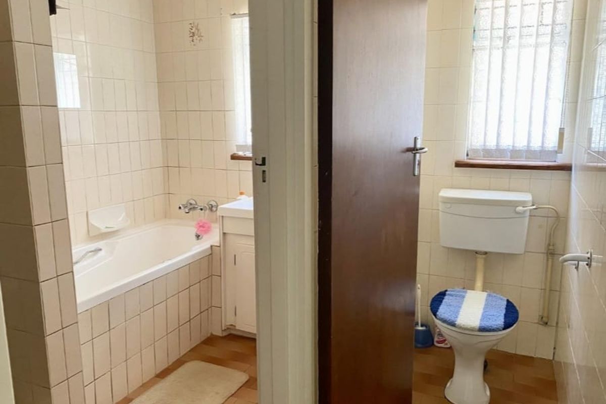 A dated bathroom featuring a built-in bathtub, vanity cabinet, and separate toilet area with original wall and floor tiling.