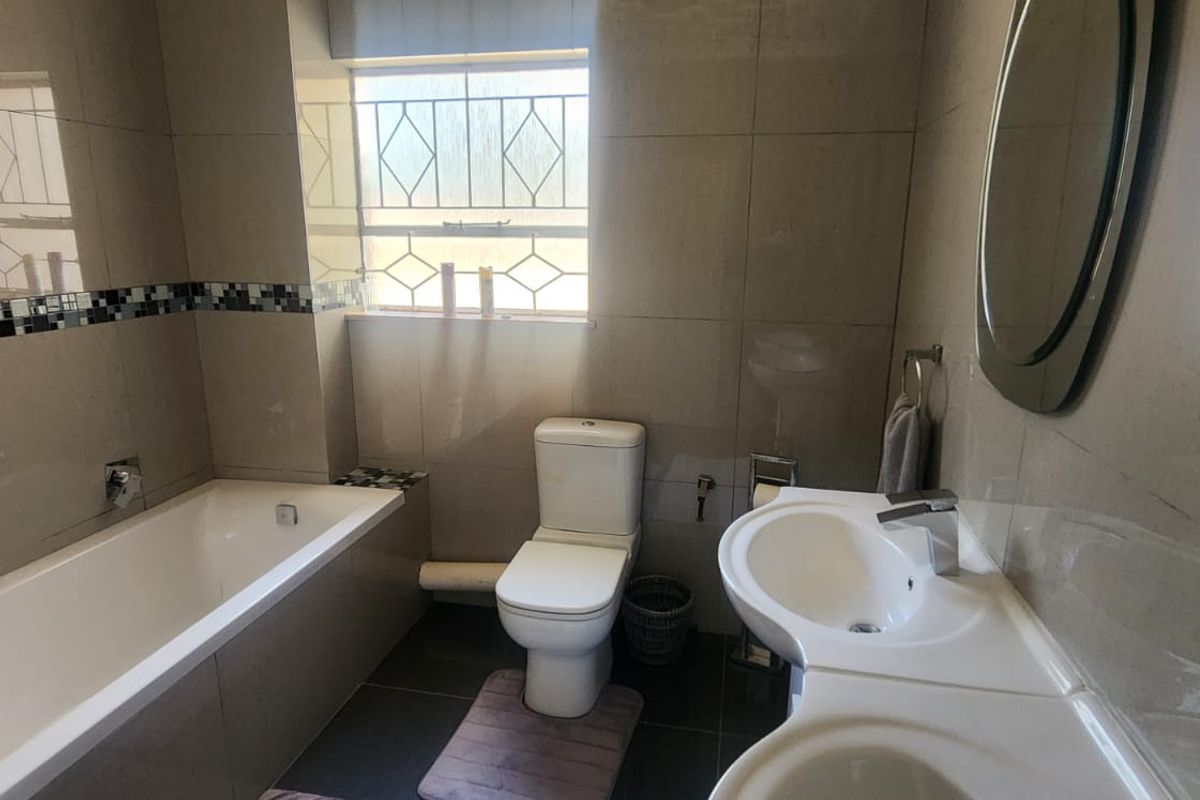 A functional bathroom featuring a built-in bathtub, a dual-basin vanity unit, and a toilet, finished with neutral wall and floor tiling.