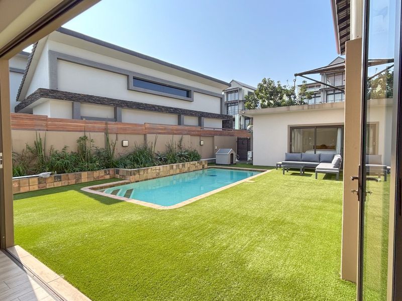A well-maintained outdoor area featuring a swimming pool, artificial lawn, and boundary wall, accessible through large sliding glass doors from the interior.