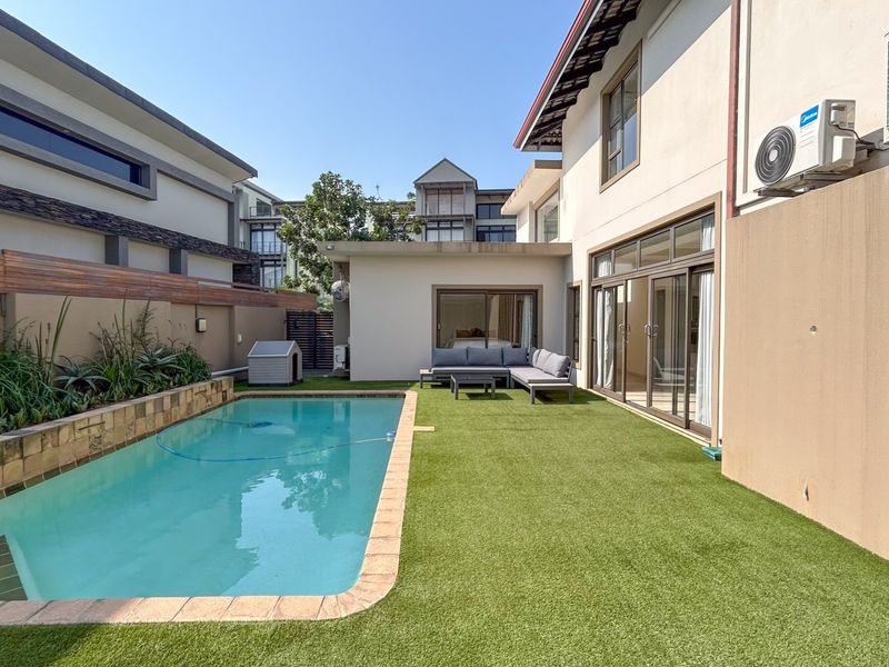 A well-maintained rear garden area featuring a swimming pool, artificial lawn, and direct access to the home via large sliding glass doors.