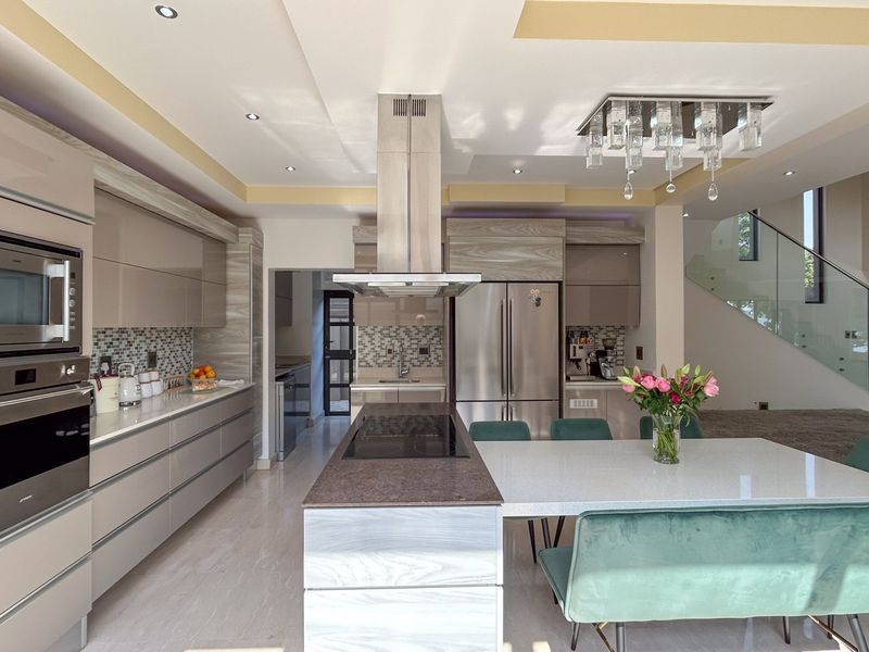 Modern open-plan kitchen featuring high-gloss cabinetry, stone countertops, a central island with integrated cooktop, and a sleek glass staircase in the background.