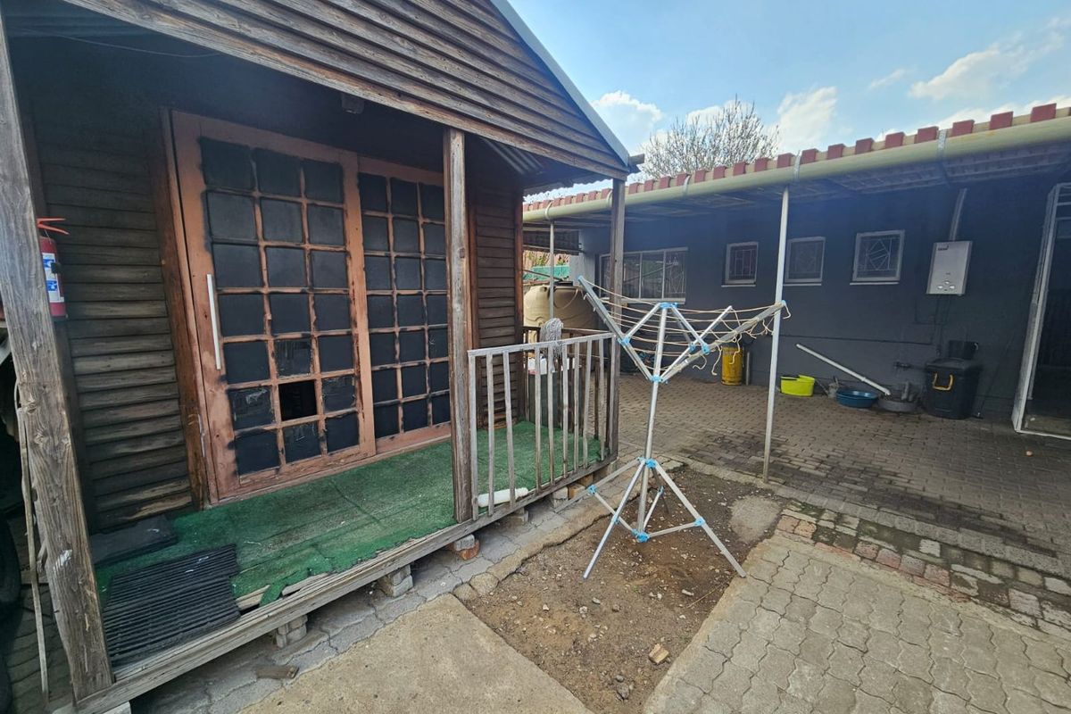 Exterior view of a weathered timber cabin featuring a small porch, adjacent to a paved courtyard area with a secondary structure.