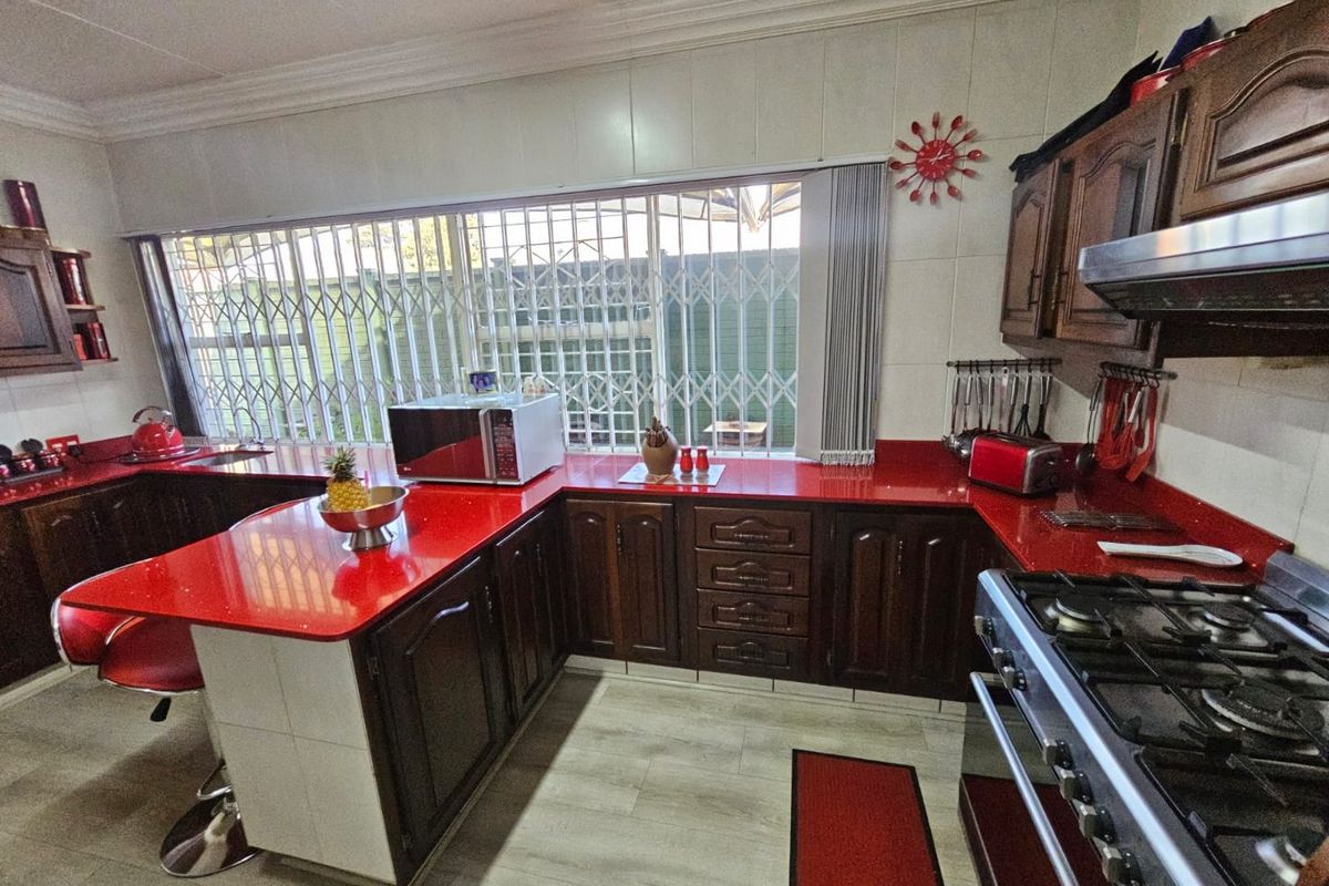 A functional kitchen featuring dark wood cabinetry, vibrant red countertops, a gas stove with extractor fan, and security bars on the window.