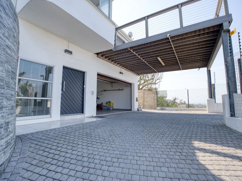 A paved driveway leading to a garage, featuring stone cladding, a covered balcony overhead, and secure perimeter fencing.