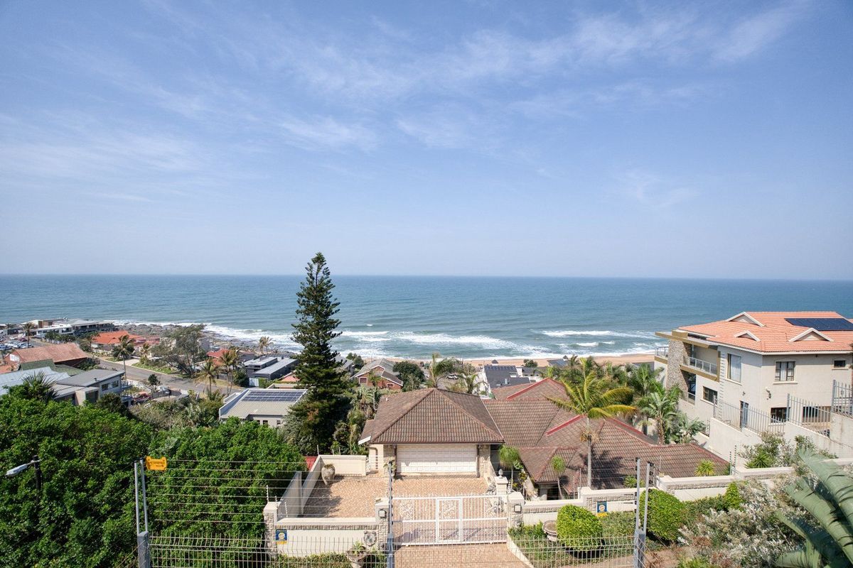 Elevated coastal view overlooking the neighborhood and ocean, featuring a residential property with a tiled roof and secure perimeter fencing.
