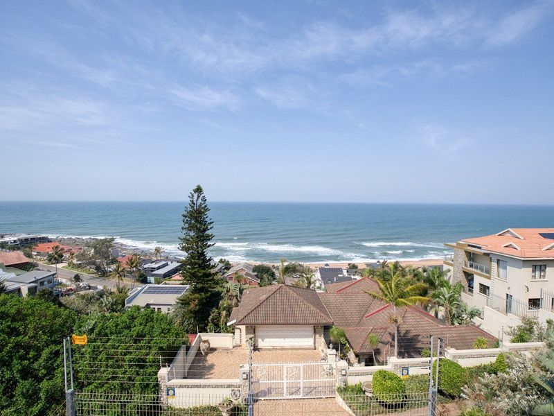 Elevated coastal view overlooking the neighborhood and ocean, featuring a residential property with a tiled roof and secure perimeter fencing.