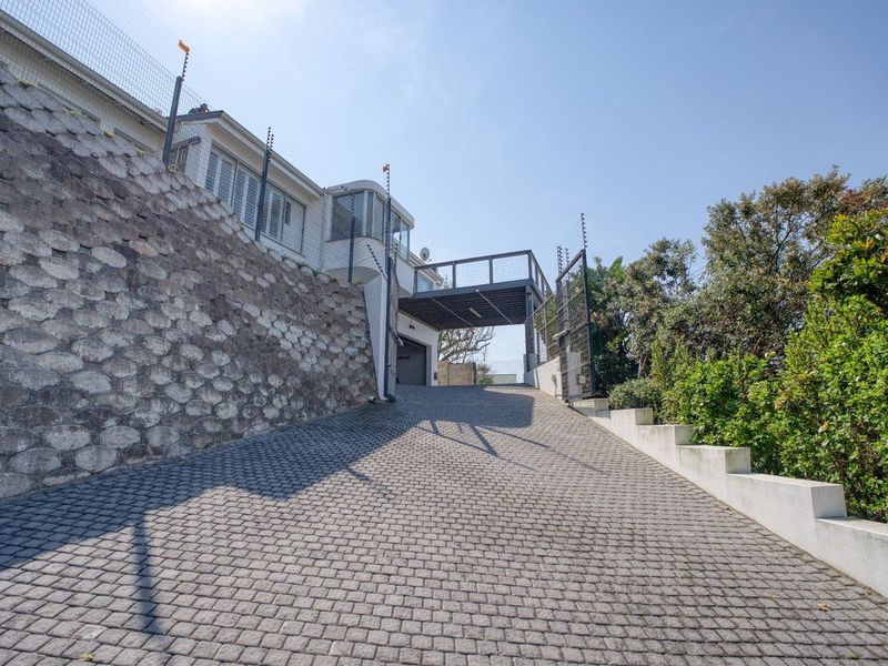 A steep cobblestone driveway leading to a garage entrance, featuring a stone retaining wall and elevated deck structure with security fencing.