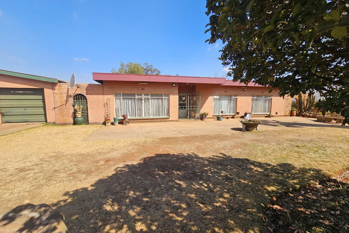 A single-story brick farmhouse featuring a paved patio area, large windows with security bars, and an attached garage, set on a spacious rural plot.