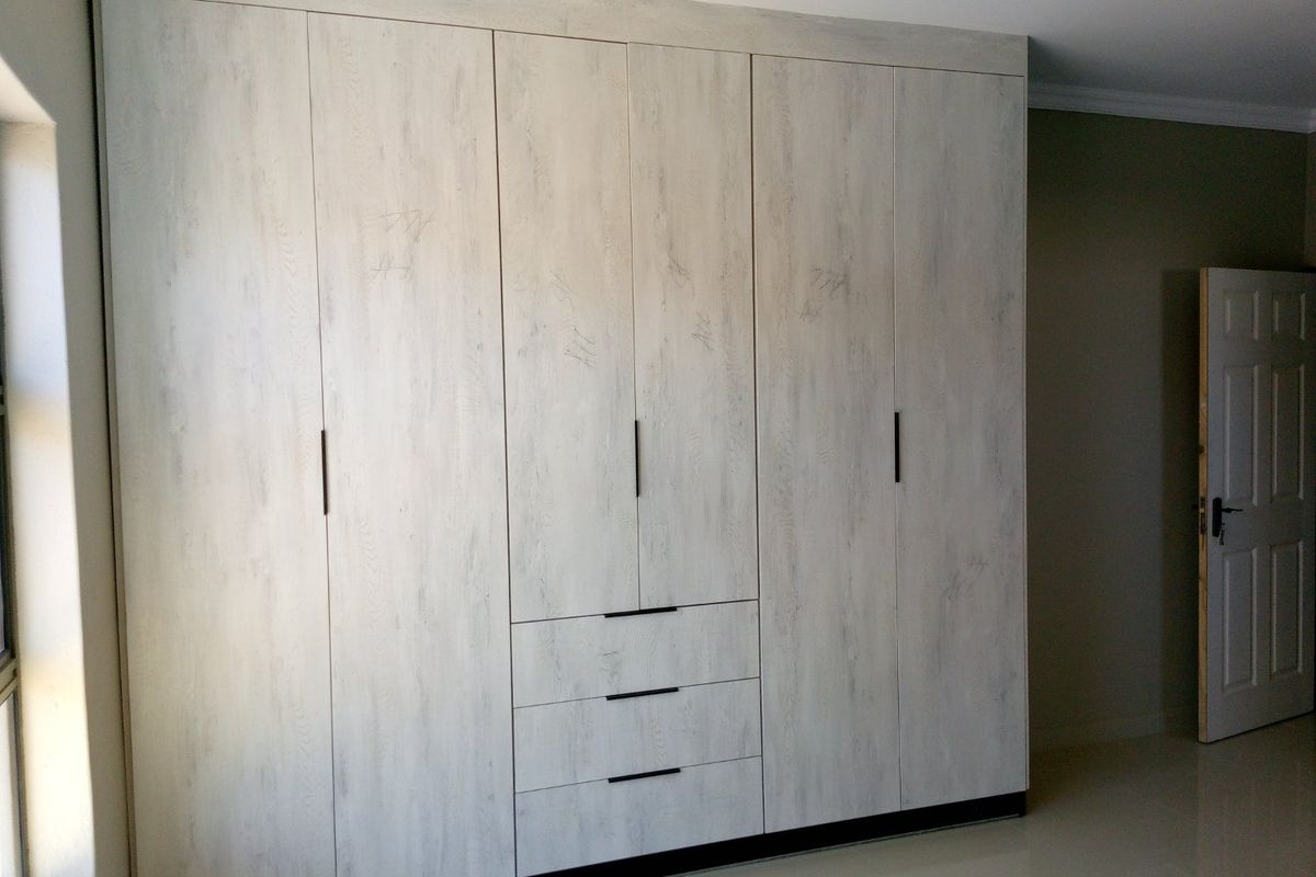 A bedroom featuring large, floor-to-ceiling built-in wardrobes with a light wood-grain finish, modern black handles, and integrated drawer storage.
