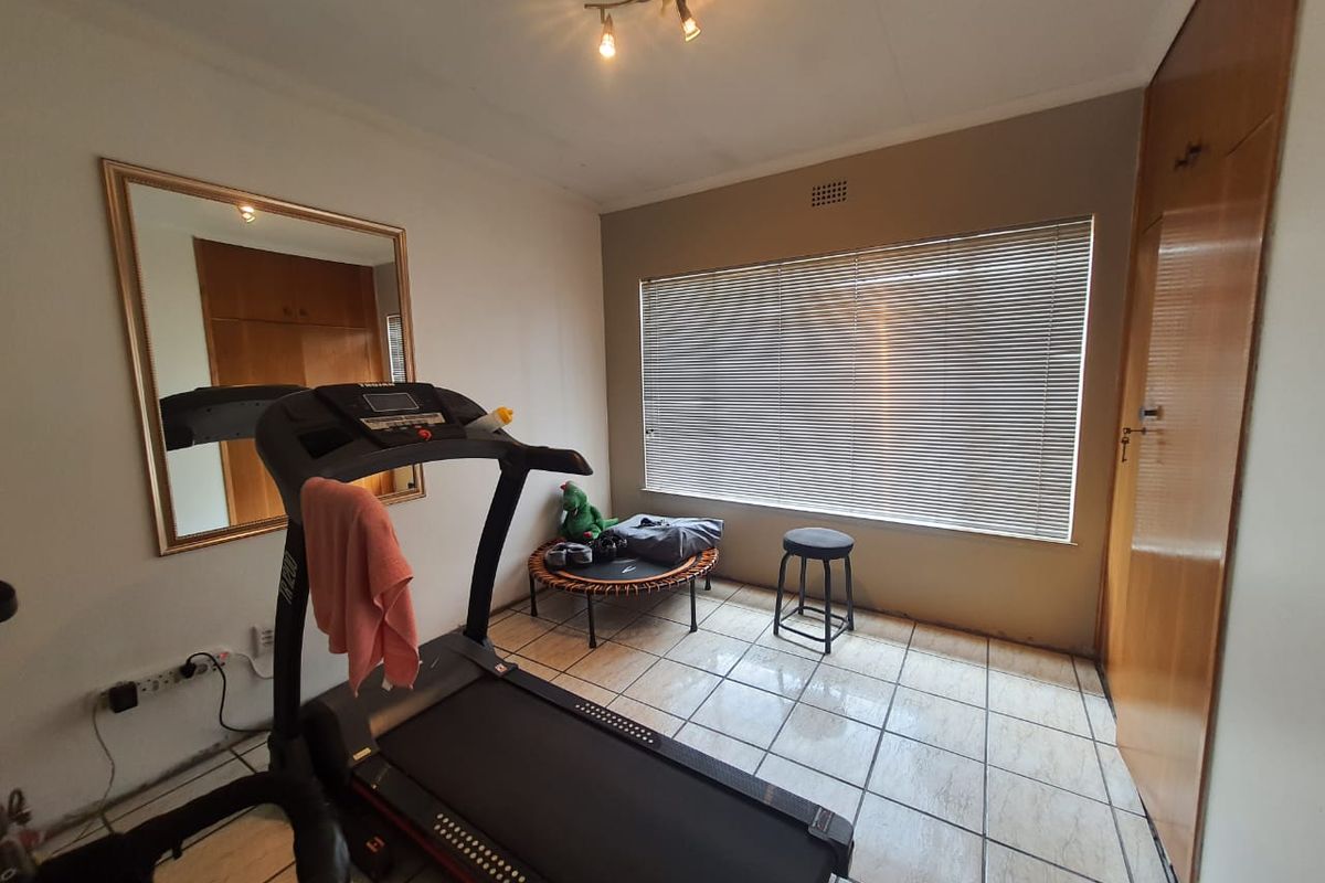 A functional bedroom featuring tiled flooring, built-in wooden cupboards, and a large window with fitted blinds, currently utilized as a home gym space.
