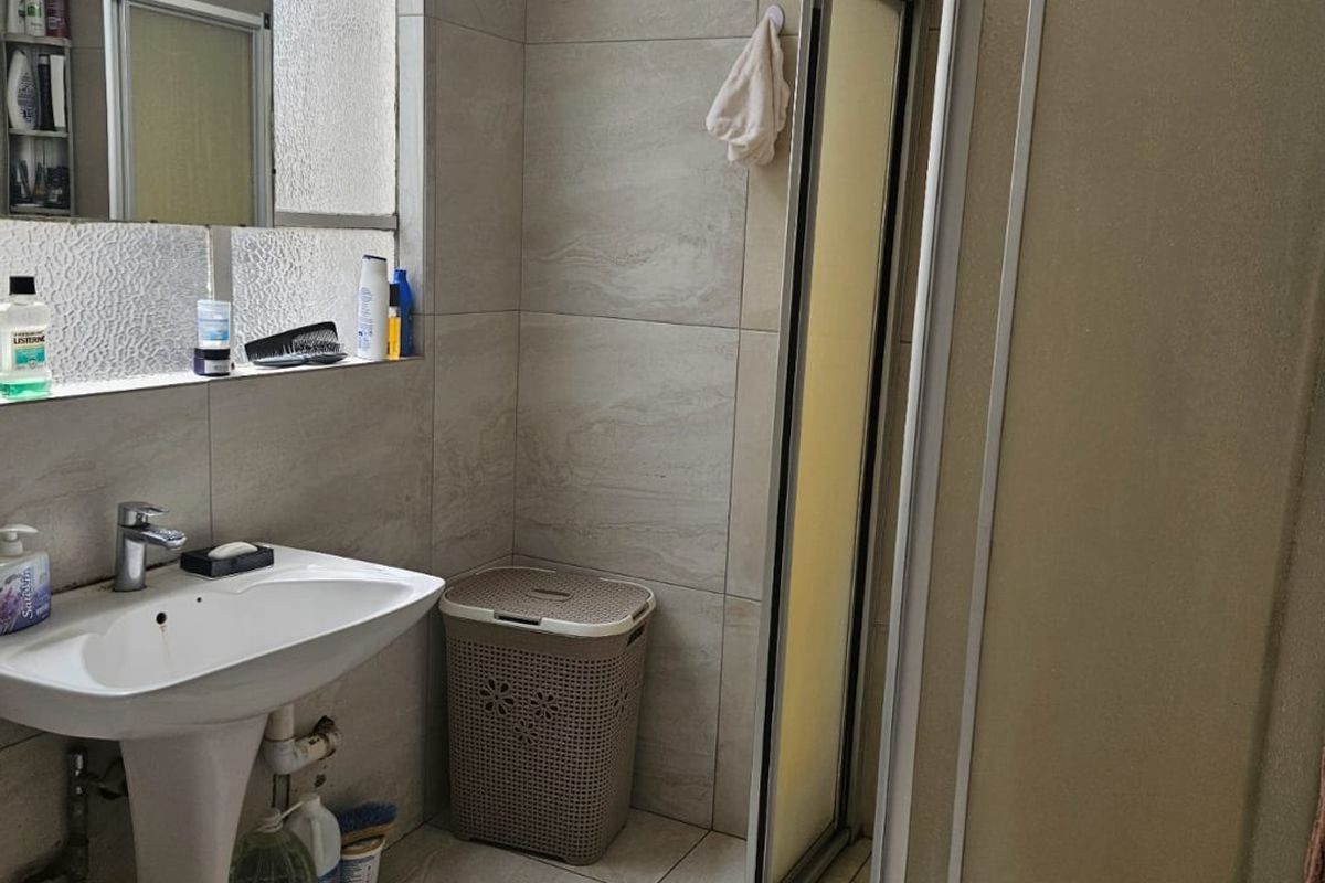 A functional bathroom featuring a pedestal basin, tiled walls and flooring, and a glass-enclosed shower unit with natural light from a high window.