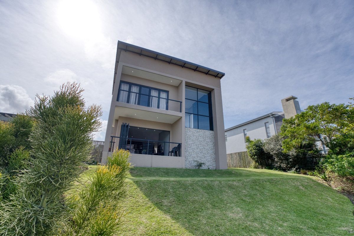 A modern two-story residential exterior featuring a rear facade with balconies, large glass windows, and stone cladding, set against a landscaped lawn.