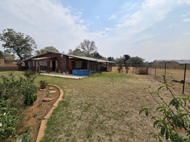 Rear view of a brick farmhouse featuring a covered patio area, expansive garden space, and perimeter fencing on a rural property.