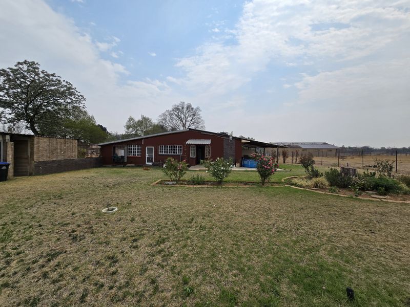 Rear view of a farm property featuring a brick dwelling with a covered patio, established garden beds, and expansive surrounding grounds.