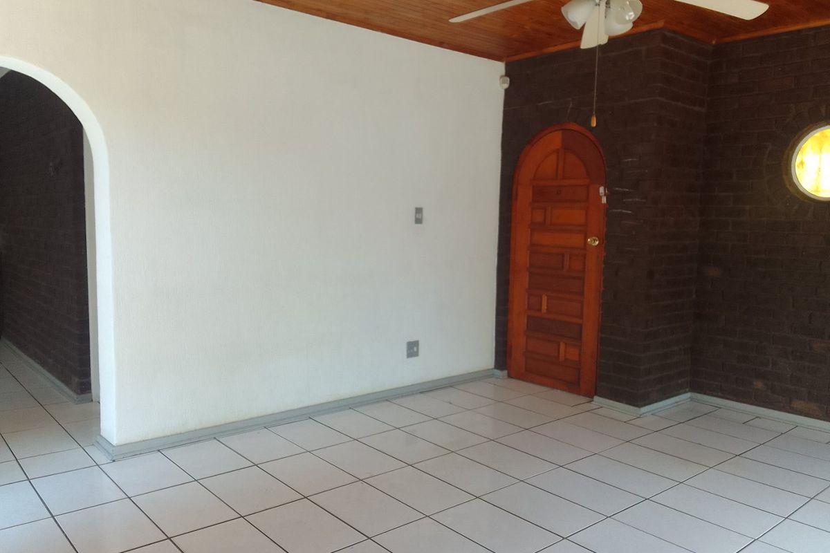 An empty living area featuring tiled floors, a timber ceiling with a fan, and a distinctive exposed brick wall with an arched timber door.