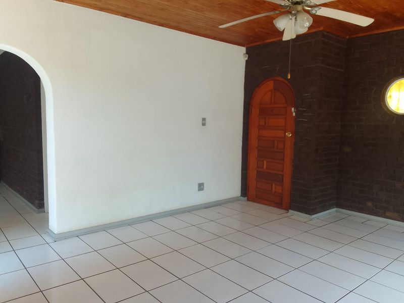 An empty living area featuring tiled floors, a timber ceiling with a fan, and a distinctive exposed brick wall with an arched timber door.