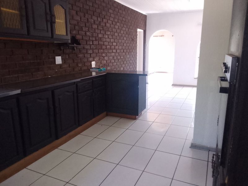 A kitchen featuring dark built-in cabinetry, an exposed brick feature wall, and white tiled flooring, leading through an archway to an adjacent living space.