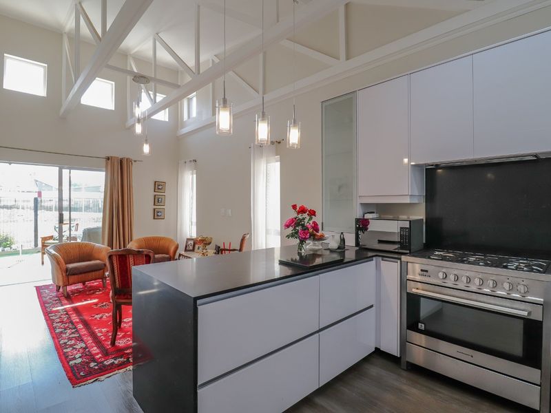 Modern open-plan kitchen and living area featuring high vaulted ceilings with exposed trusses, sleek cabinetry, stone countertops, and direct access to a patio.