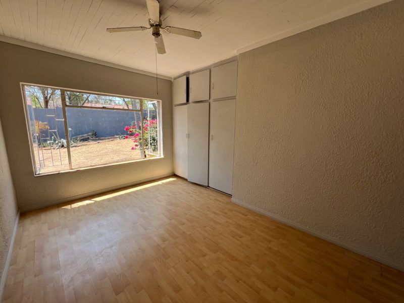 A bright, empty bedroom featuring built-in cupboards, a ceiling fan, and large windows overlooking the rear garden.