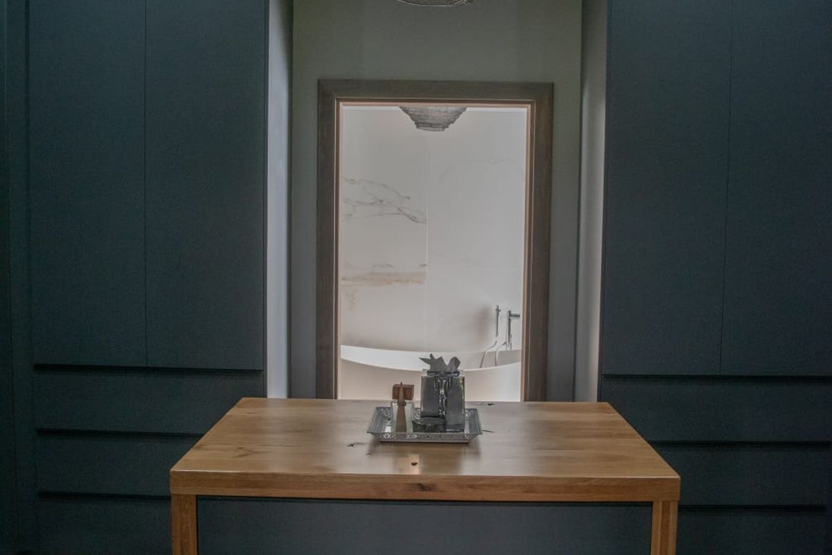 A modern walk-in dressing room featuring floor-to-ceiling dark cabinetry, a central wooden island unit, and a pendant light, leading into an adjacent bathroom.