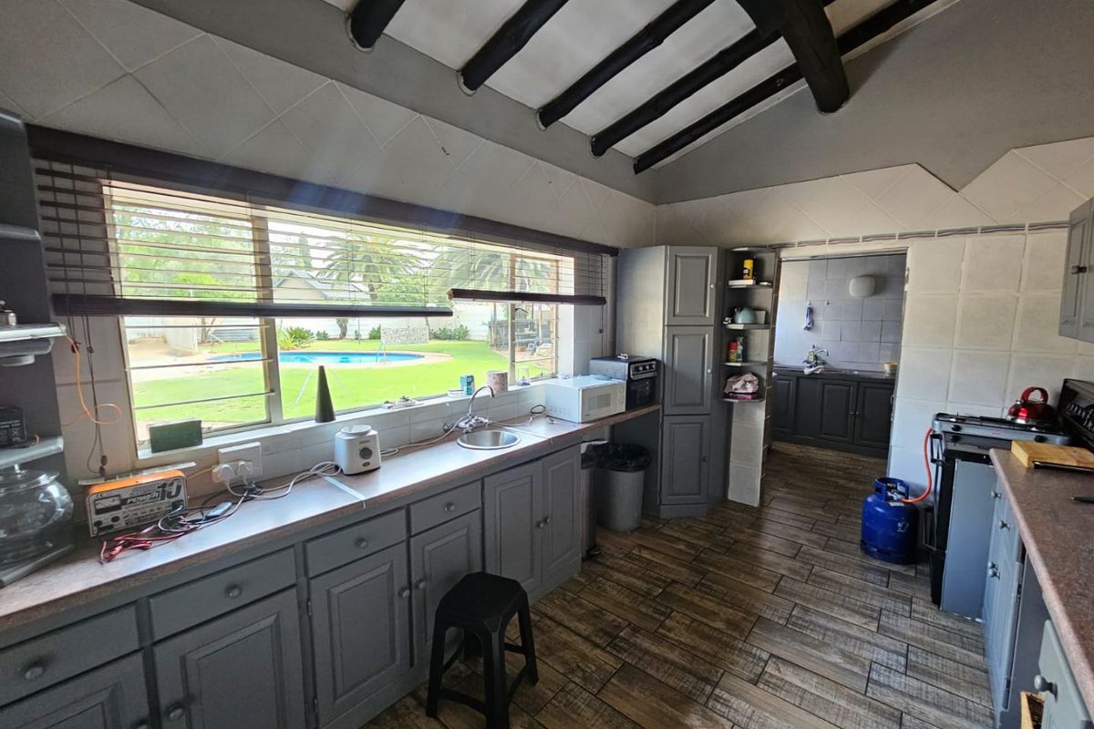 A functional kitchen featuring grey built-in cabinetry, laminate countertops, tiled flooring, and exposed ceiling beams, with a view overlooking the garden and pool area.