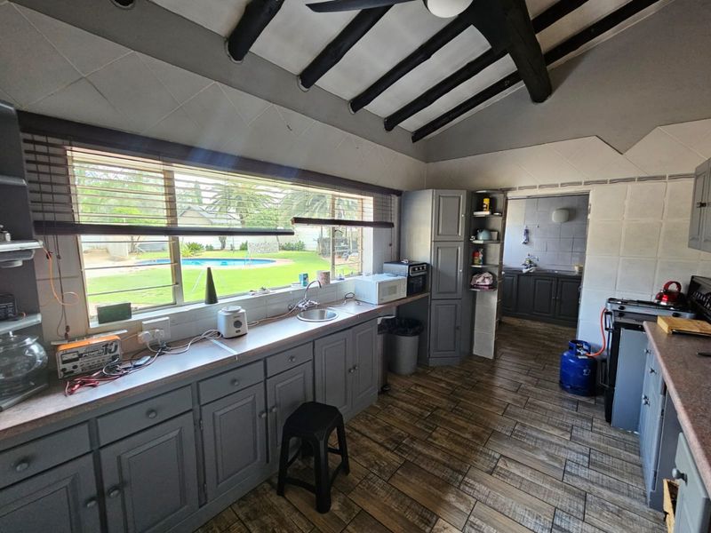 A functional kitchen featuring grey built-in cabinetry, laminate countertops, tiled flooring, and exposed ceiling beams, with a view overlooking the garden and pool area.