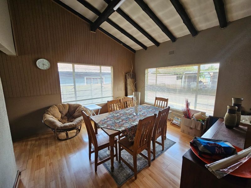 Dining room featuring exposed timber ceiling beams, laminate flooring, and large windows with vertical blinds, offering a rustic aesthetic with potential for modernization.