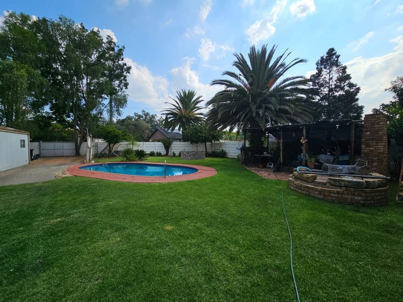 Spacious backyard featuring a swimming pool with brick coping, a covered patio area, and a built-in brick braai, set within a landscaped garden.