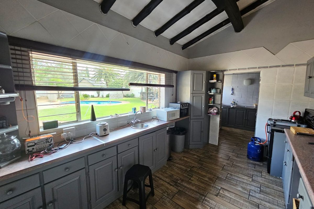 A functional kitchen featuring grey cabinetry, tiled flooring, and exposed ceiling beams, with a view overlooking the garden and swimming pool area.