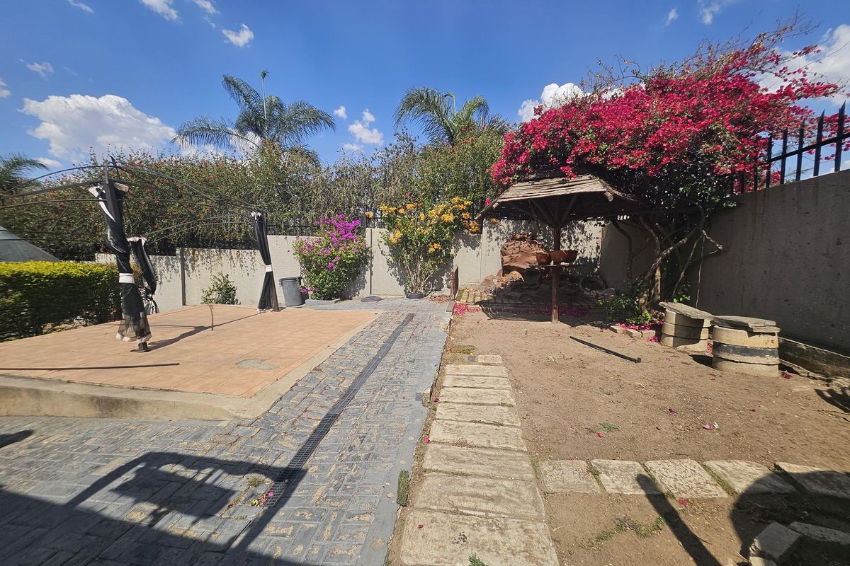Rear garden area featuring a paved patio, drainage channel, stone pathway, and a small thatched gazebo against a perimeter wall.