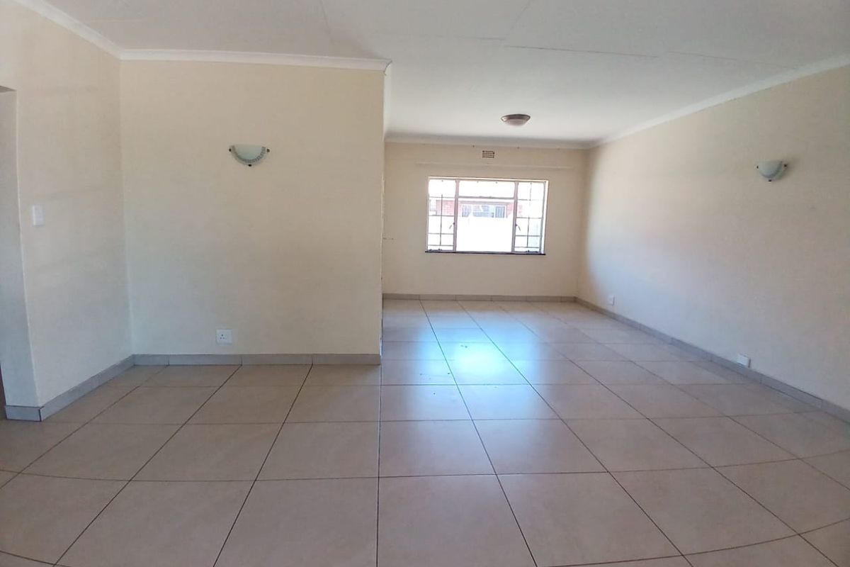 A spacious, empty living area featuring neutral tiled flooring, wall-mounted light fixtures, and a large window providing natural light.