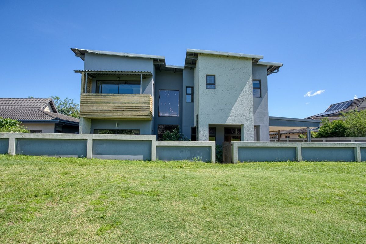 Rear view of a modern two-story house featuring a balcony with timber cladding, large windows, and a surrounding boundary wall set on a lawn.