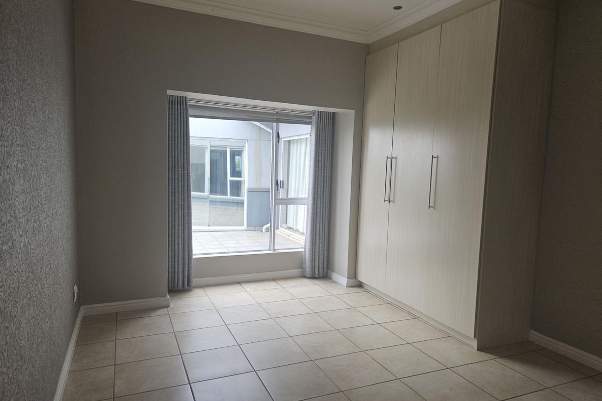 A bright, empty bedroom featuring neutral tiled flooring, built-in cupboards, and a large window providing natural light and access to an outdoor area.