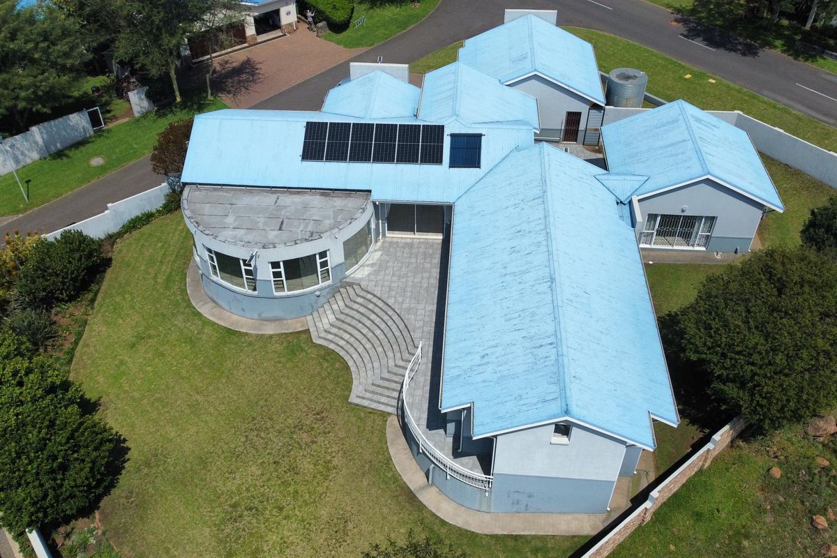 Aerial view of a residential property featuring a multi-wing layout, blue metal roofing with integrated solar panels, and a paved rear patio area.