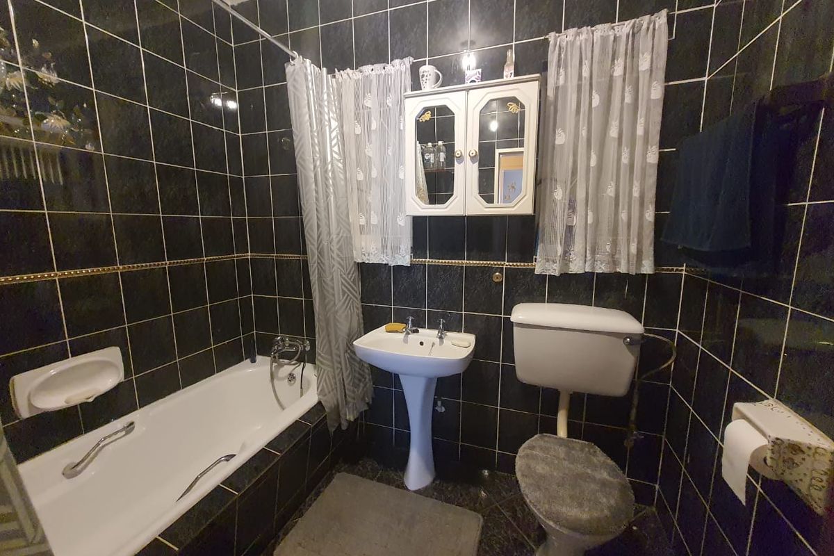 A dated bathroom featuring dark ceramic wall tiles, a white pedestal basin, a standard bathtub with shower mixer, and a wall-mounted medicine cabinet.
