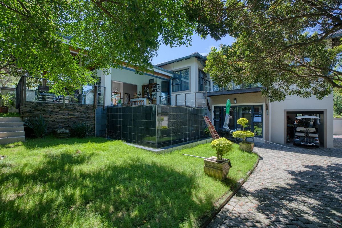 Rear view of the property featuring a landscaped garden, stone retaining wall, tiled water feature, raised patio with glass balustrade, and paved driveway.
