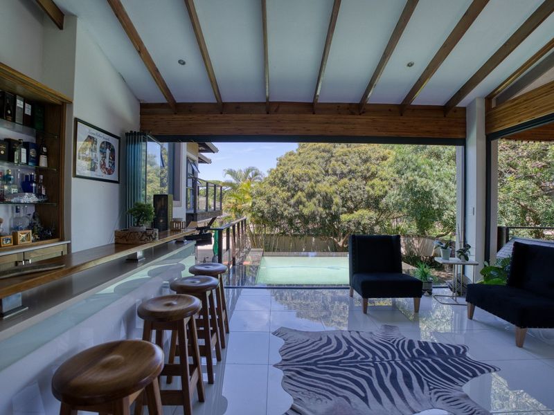 Modern indoor bar area featuring built-in shelving, exposed timber beams, and large sliding doors opening to a swimming pool and garden view.