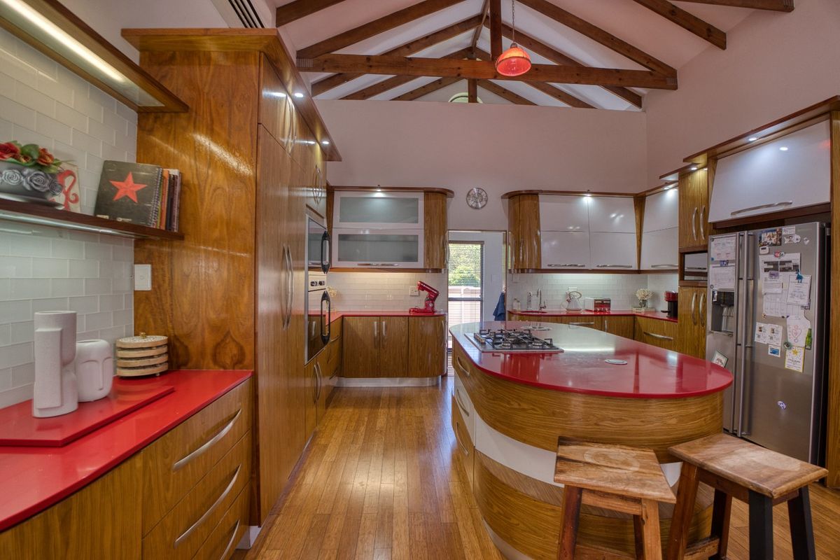 Spacious kitchen featuring custom wooden cabinetry, vibrant red countertops, a central island with gas hob, and striking exposed timber roof trusses.