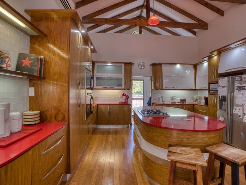 Spacious kitchen featuring custom wooden cabinetry, vibrant red countertops, a central island with gas hob, and striking exposed timber roof trusses.
