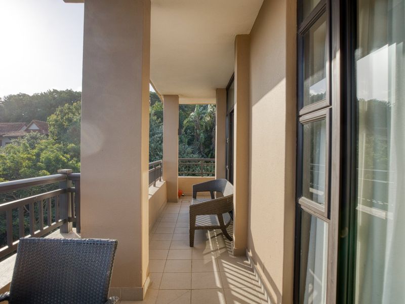 A covered balcony featuring tiled flooring, metal balustrades, and sliding glass door access, offering a pleasant outdoor space with views of surrounding greenery.