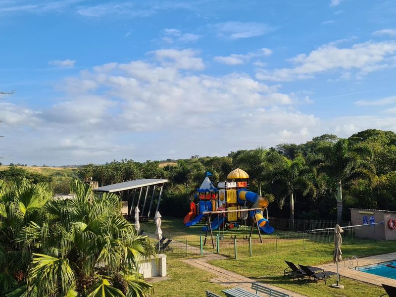 Outdoor recreational area featuring a children's playground, swimming pool, and covered seating space set within a landscaped garden environment.