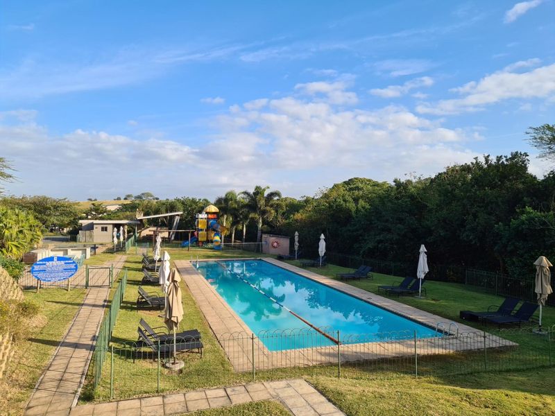 A well-maintained outdoor swimming pool area featuring a paved surround, perimeter fencing, and adjacent landscaped garden with playground facilities.
